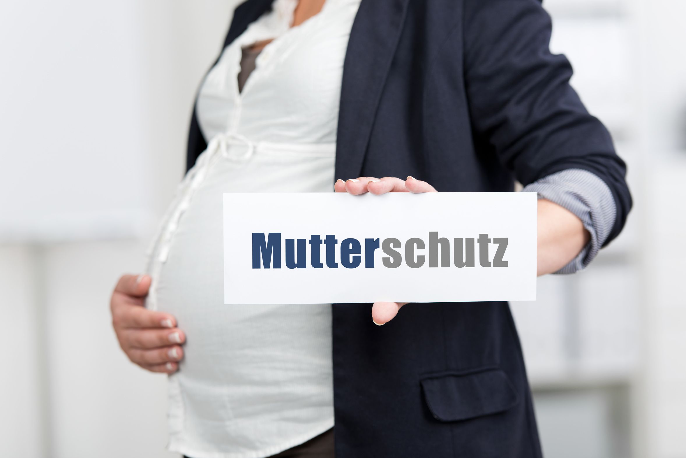 Schwangere Frau in weißer Bluse und dunklem Blazer hält sich mit rechter Hand den Bauch und in der Linken ein Schild mit der Aufschrift Mutterschutz.