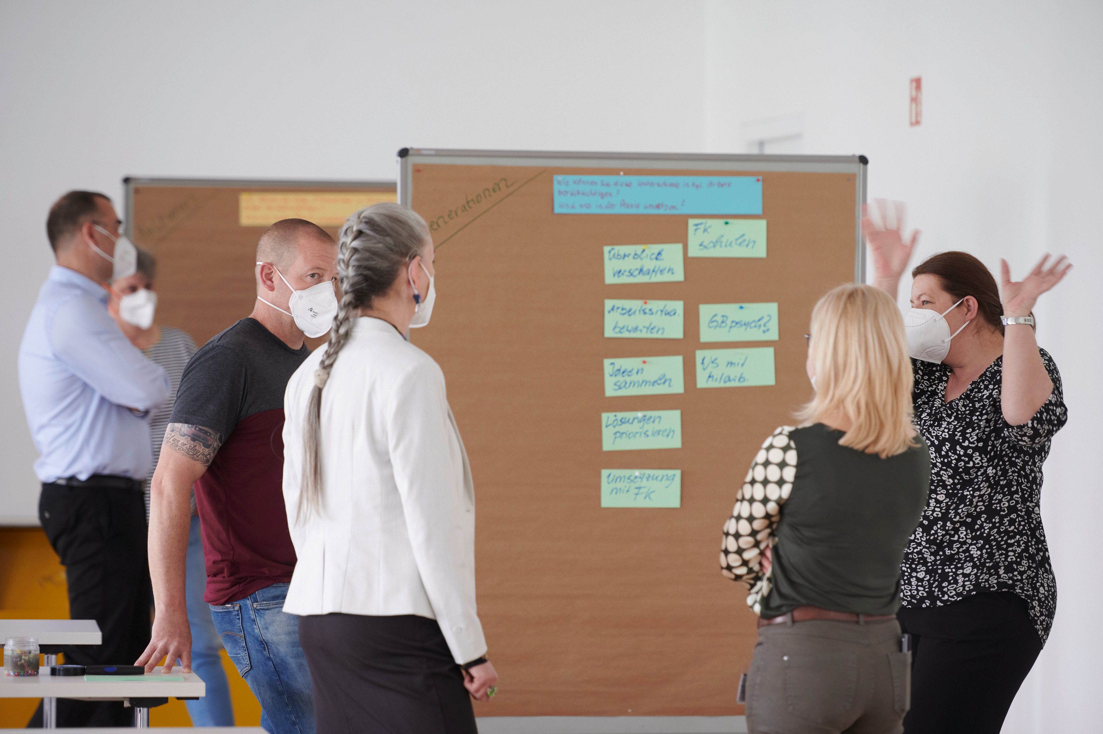 Fünf Teilnehmende stehen an einer Pinwand in einem Workshop. Alle tragen einen Mund-Nasen-Schutz.