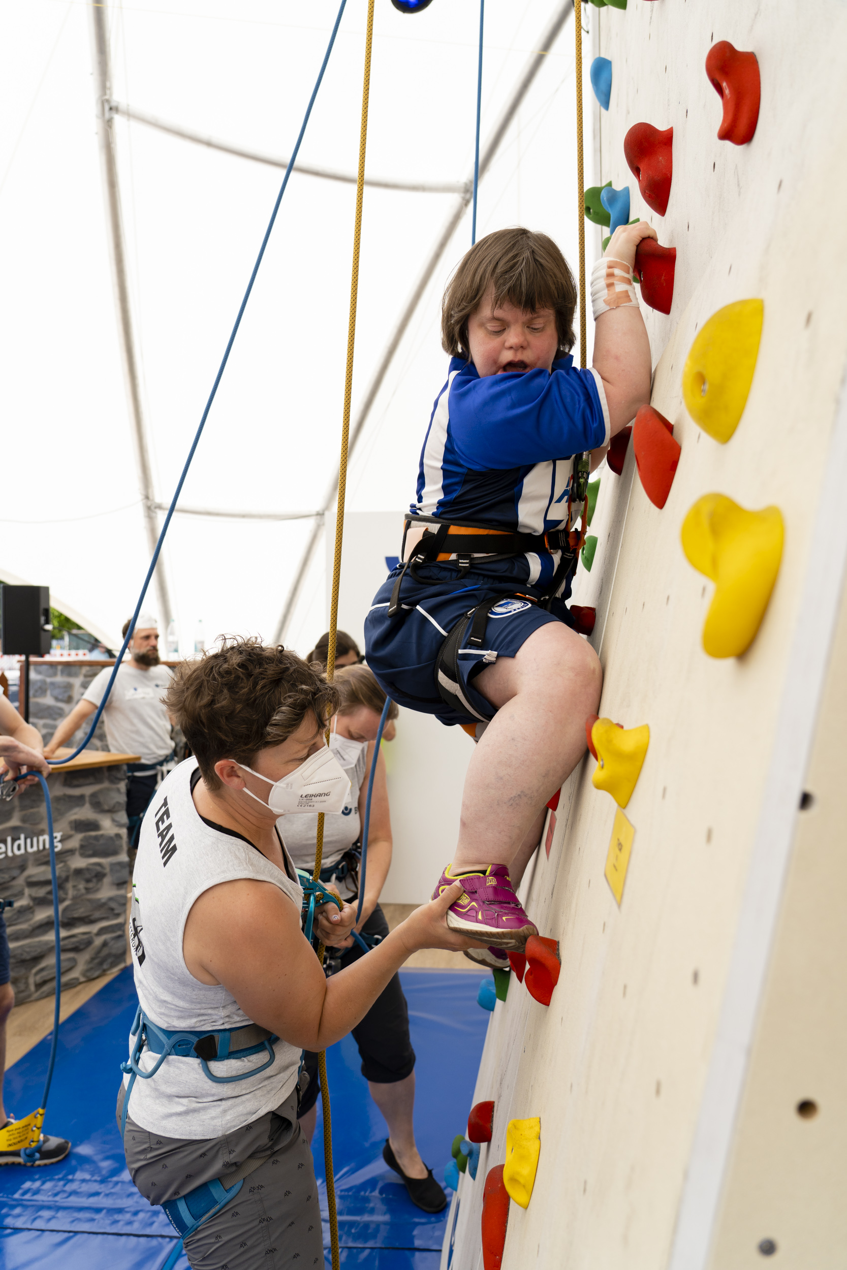 Besucherin erklimmt freudig inklusiven Kletterturm im BGW-Pavillon, BGW-Volunteers helfen ihr dabei