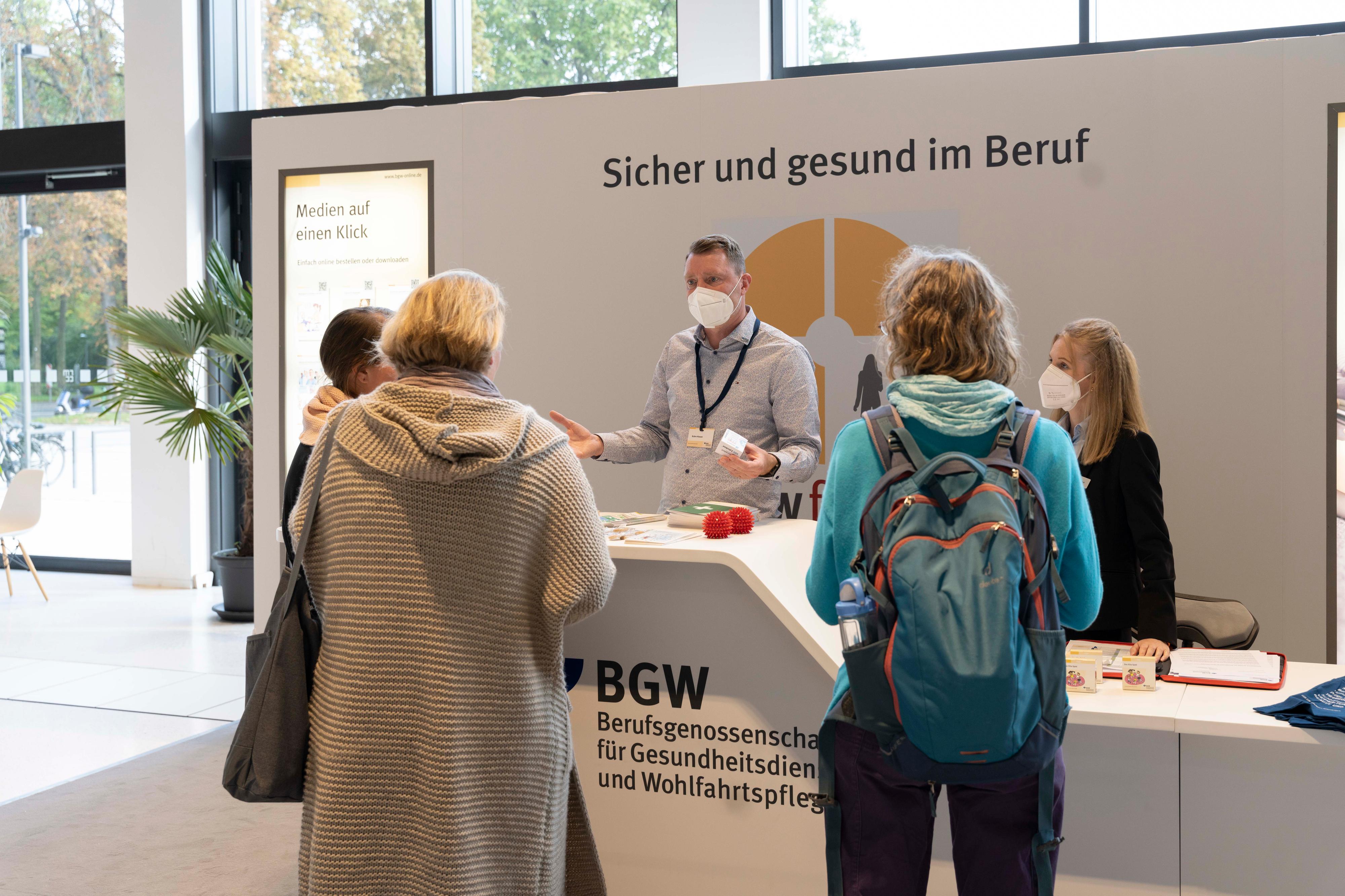 Mehrere Personen stehen am einen Messestand, ein Mann mit Maske hinter dem Tresen spricht mit ihnen, im Hintergrund steht an der Rückwand der Text "Sicher und gesund im Beruf"