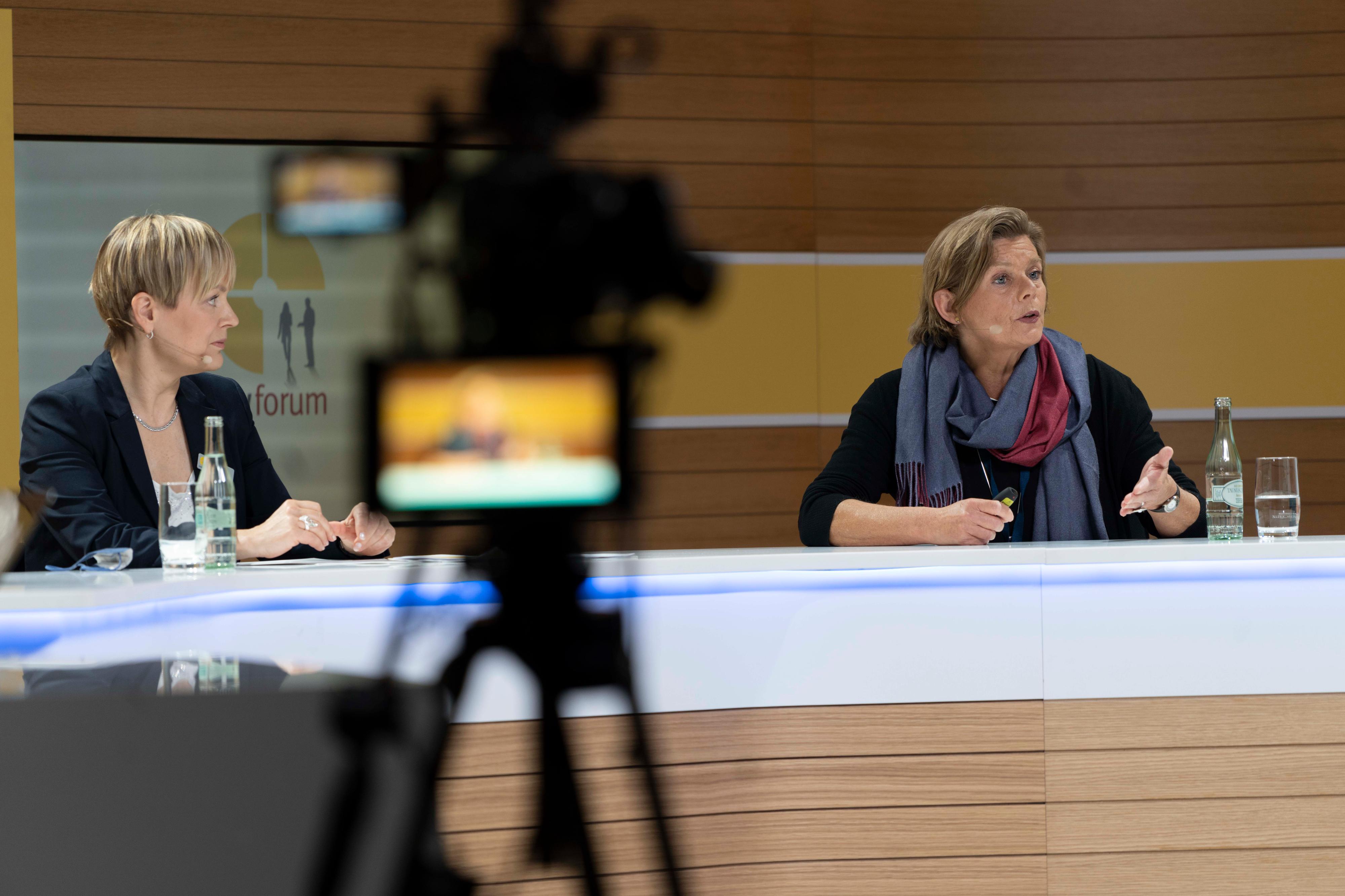 Zwei Frauen sitzen an einem Tisch wie in einem Fernsehstudio, im Bildvordergrund eine verschwommene Kamera, durch die sie gefilmt werden