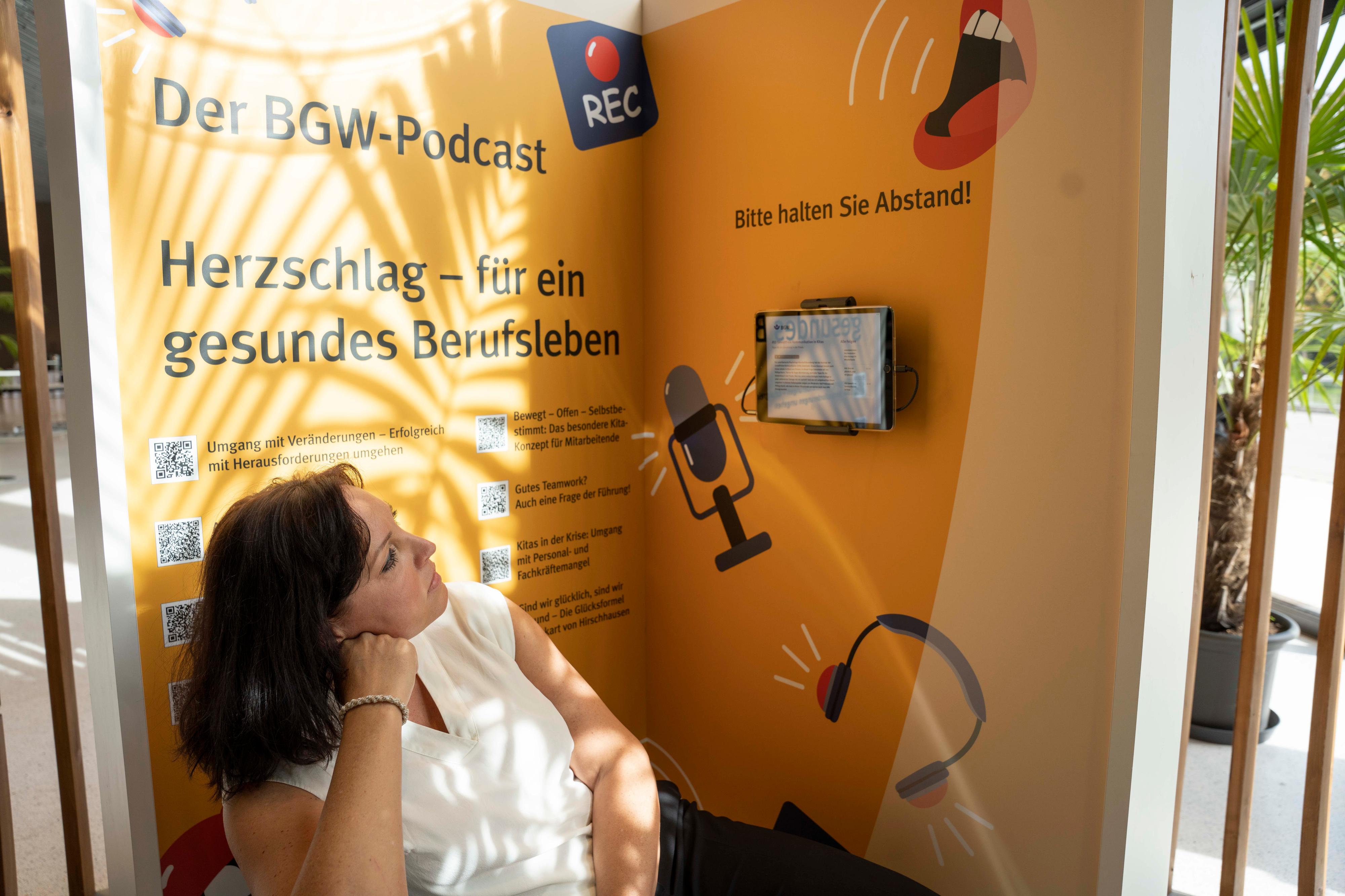 Eine Frau sitzt vor einer Wand mit dem Text "Der BGW-Podcast" und "Herzschlag - für ein gesundes Berufsleben".