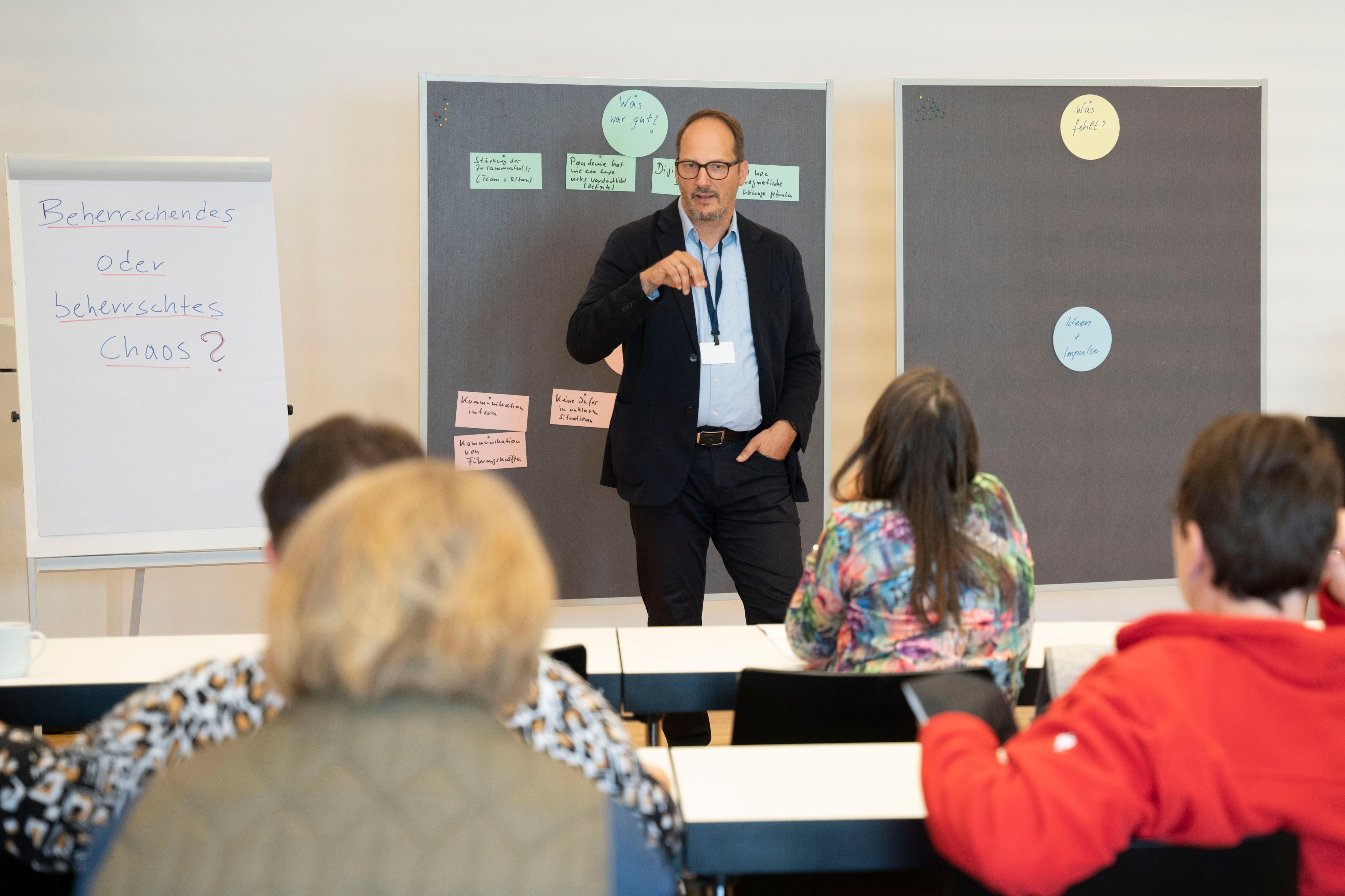 Ein Mann steht in einem Seminarraum vor Teilnehmenden. Hinter ihm befinden sich zwei Pinwände mit Karten, neben ihm steht ein Flipchart mit der Aufschrift: "Beherrschendes oder beherrschtes Chaos?"