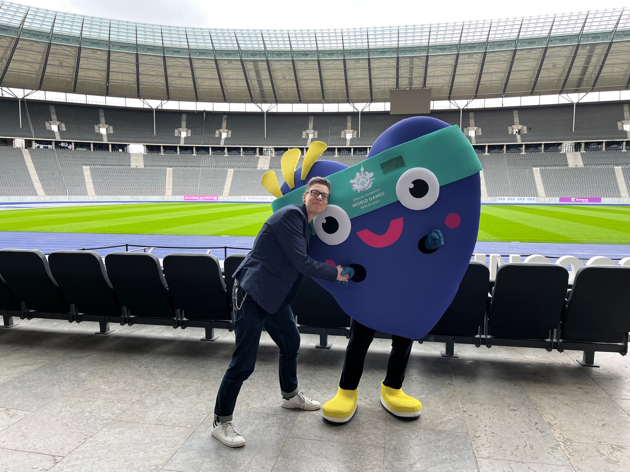 Eine Frau steht im Olympiastadion Berlin neben dem Maskottchen Unity - ein verkleideter Mensch -und gibt ihm die Hand.