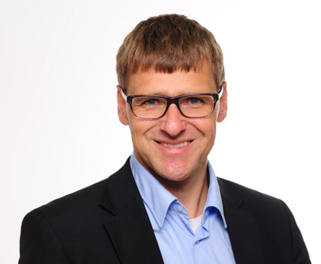 Andreas Kummer Mann mit blonden Haaren, schwarzer Brille, schwarzen Blazer und blauen Hemd lächelt in die Kamera.