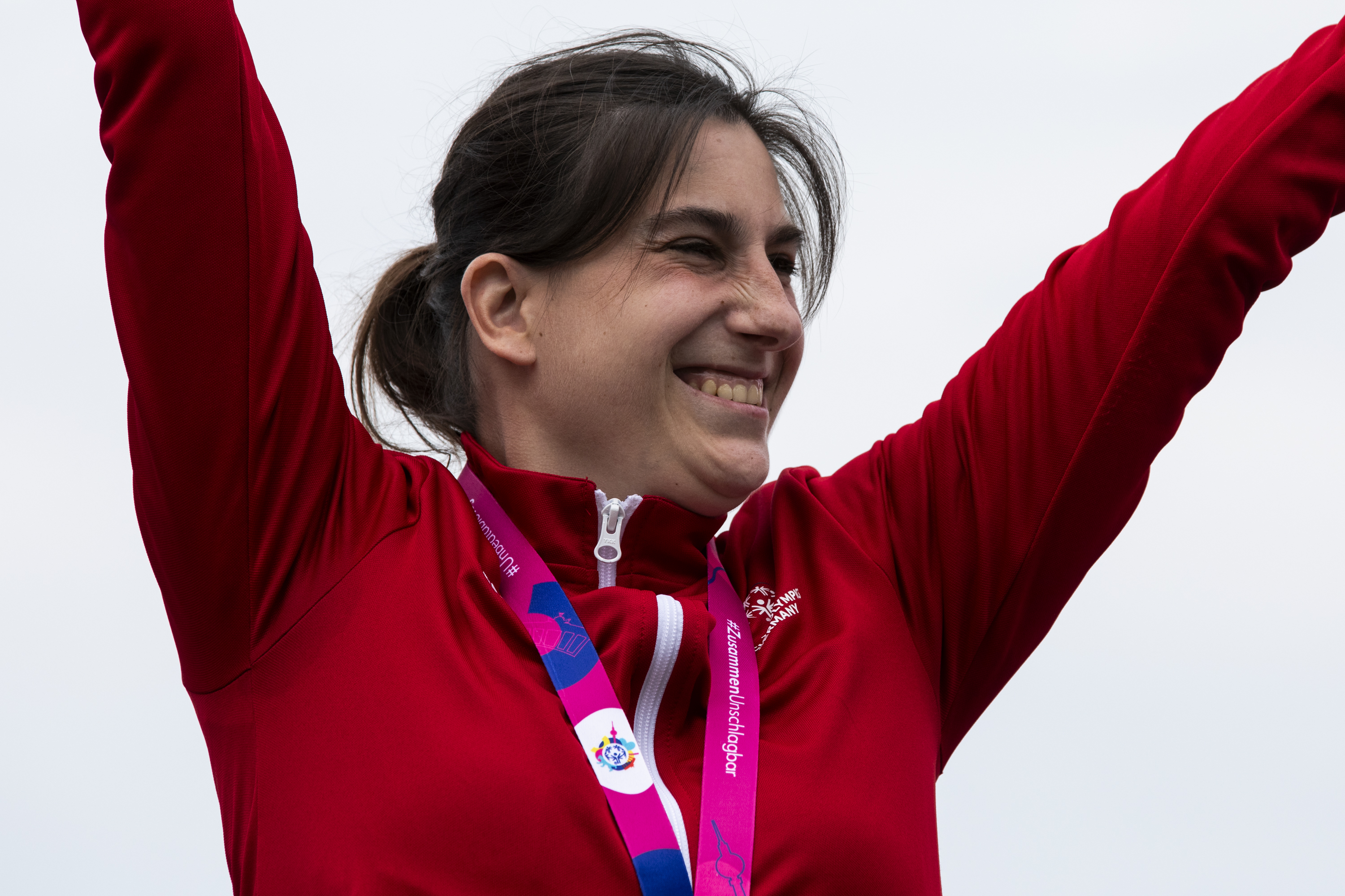 Patrizia Spaulding mit Goldmedaille in roter Trainingsjacke reißt beide Arme hoch und lacht.