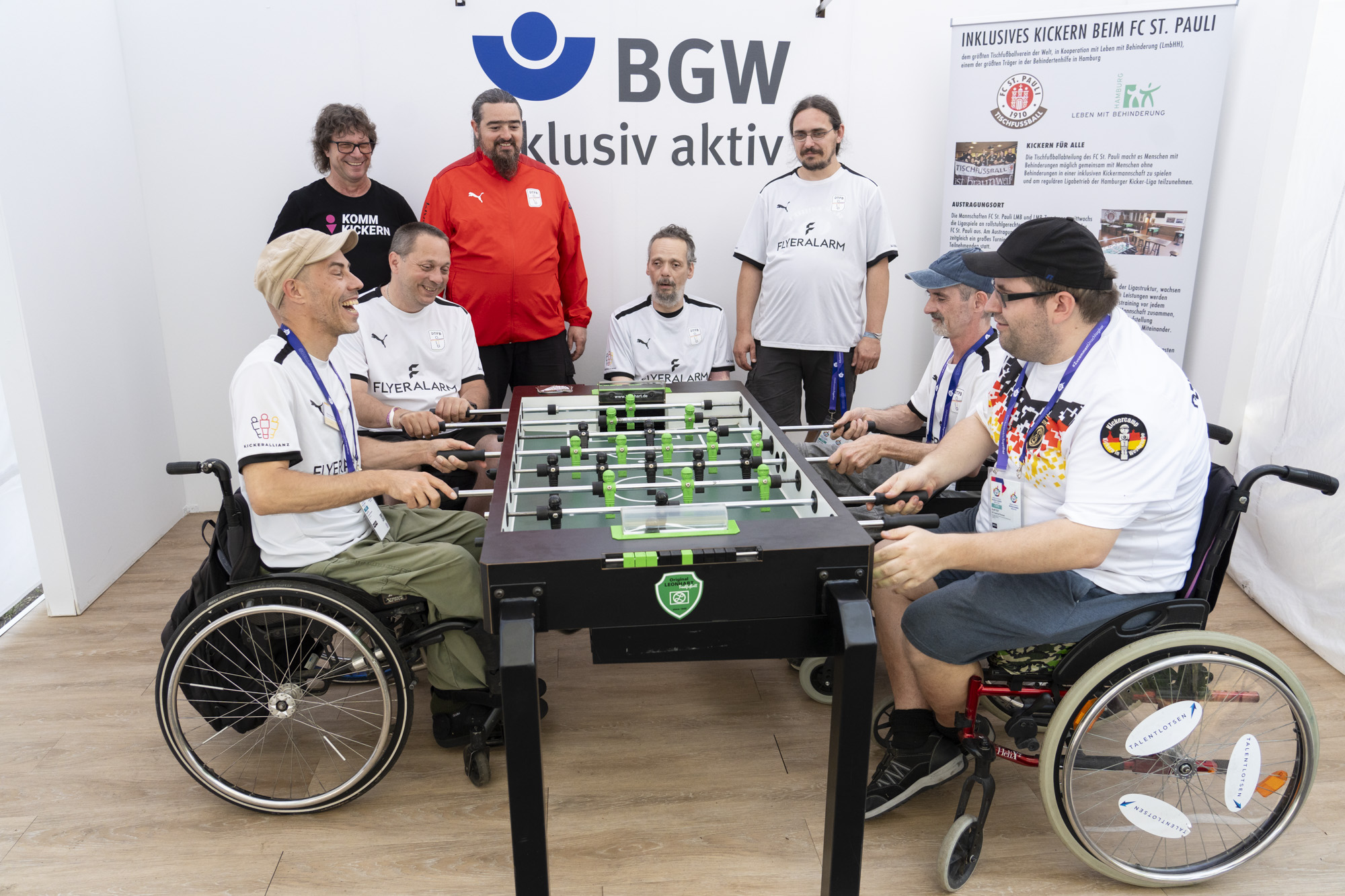 Vier Männer im Rollstuhl spielen an einem Tischfußball-Tisch. Im Hintergrund 4 weitere Männer in Trikots und eine Infotafel "Inklusives Kickern beim FC St. Pauli". An der Wand daneben der Schriftzug "BGW inklusiv aktiv"