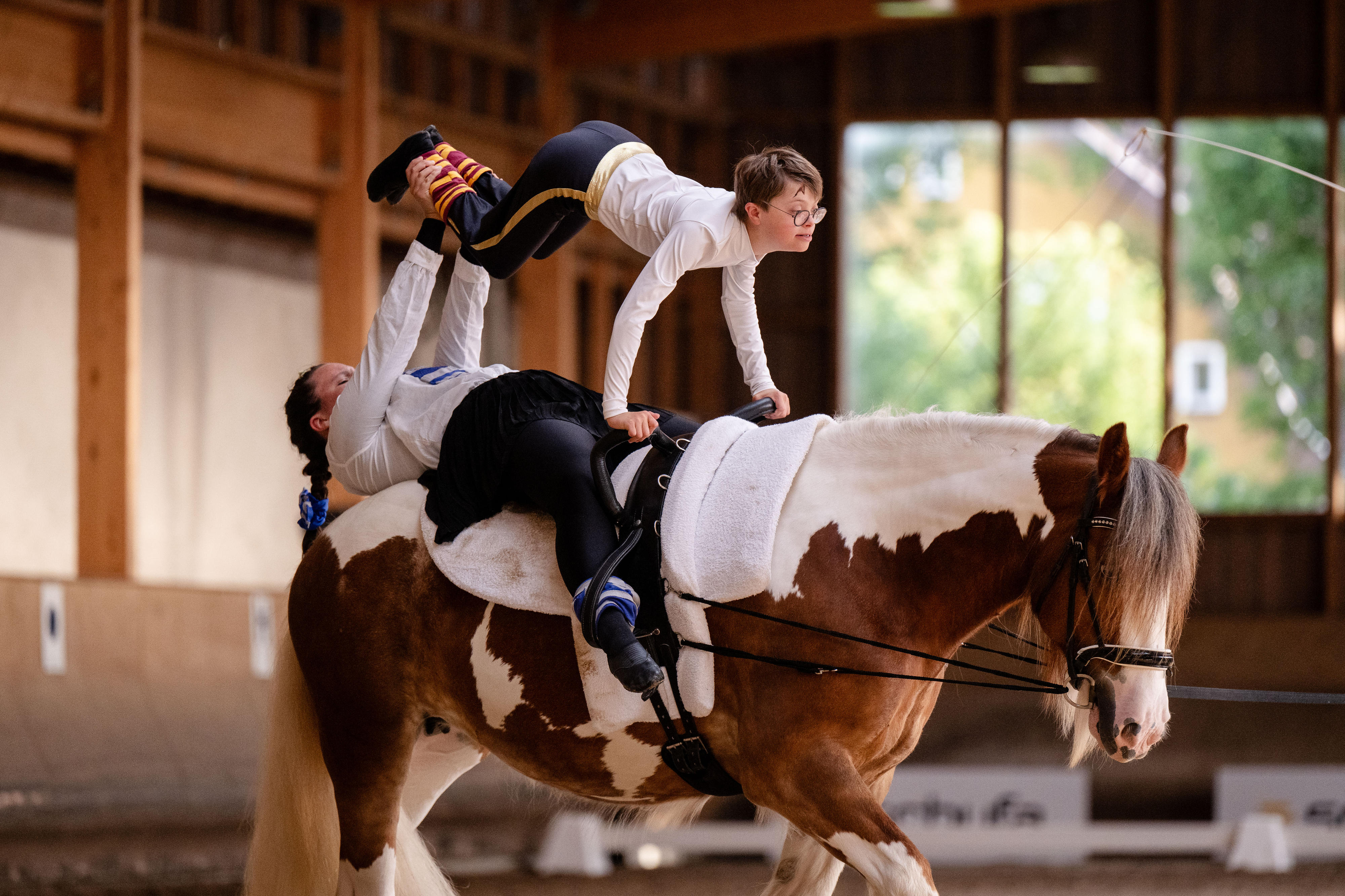 Ein Mädchen liegt auf einem Pferd und hält dabei über sich einen Jungen an den Füßen hoch. Er stützt sich mit den Händen am Sattel ab.