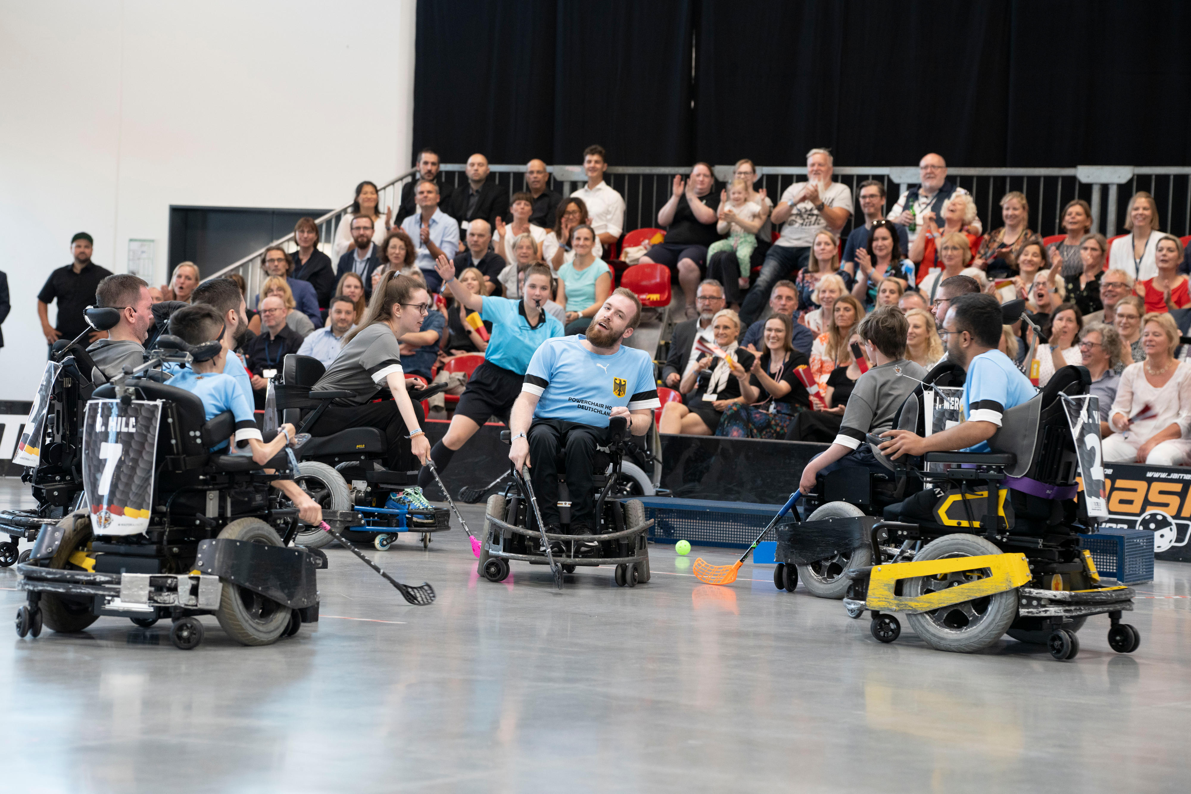 Hockey-Spielerinnen und -Spieler in elektrischen Rollstühlen in einer Sporthalle, im Hintergrund applaudiert das Publikum.