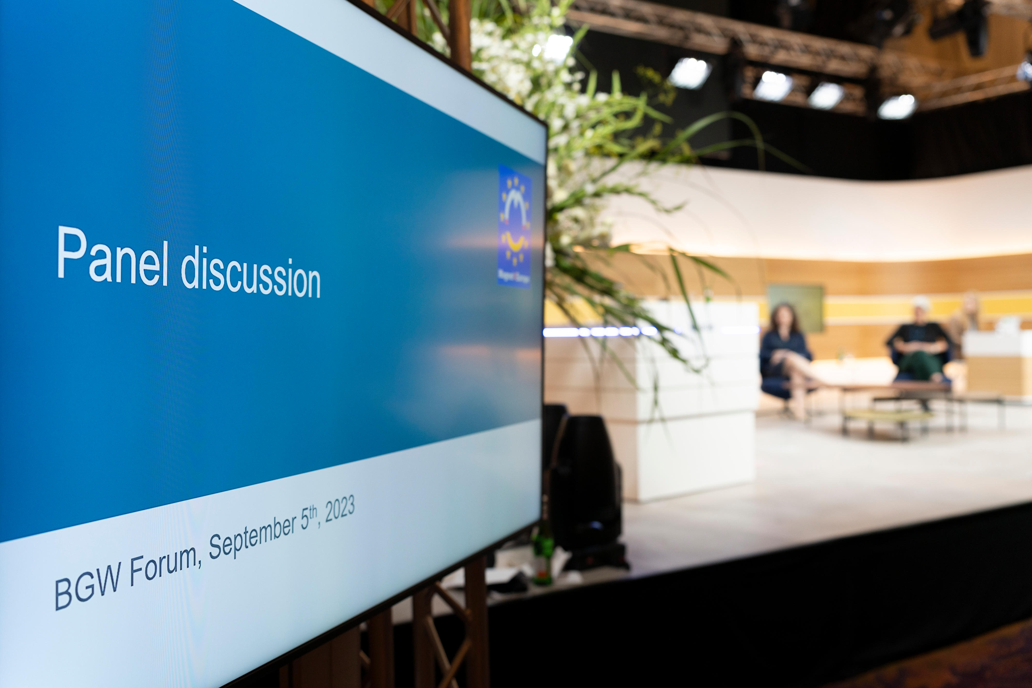 Auf einem Bildschirm im Vordergrund steht "Panel discussion", im unscharfen Hintergrund sitzen 2 Menschen auf einer beleuchteten Bühne