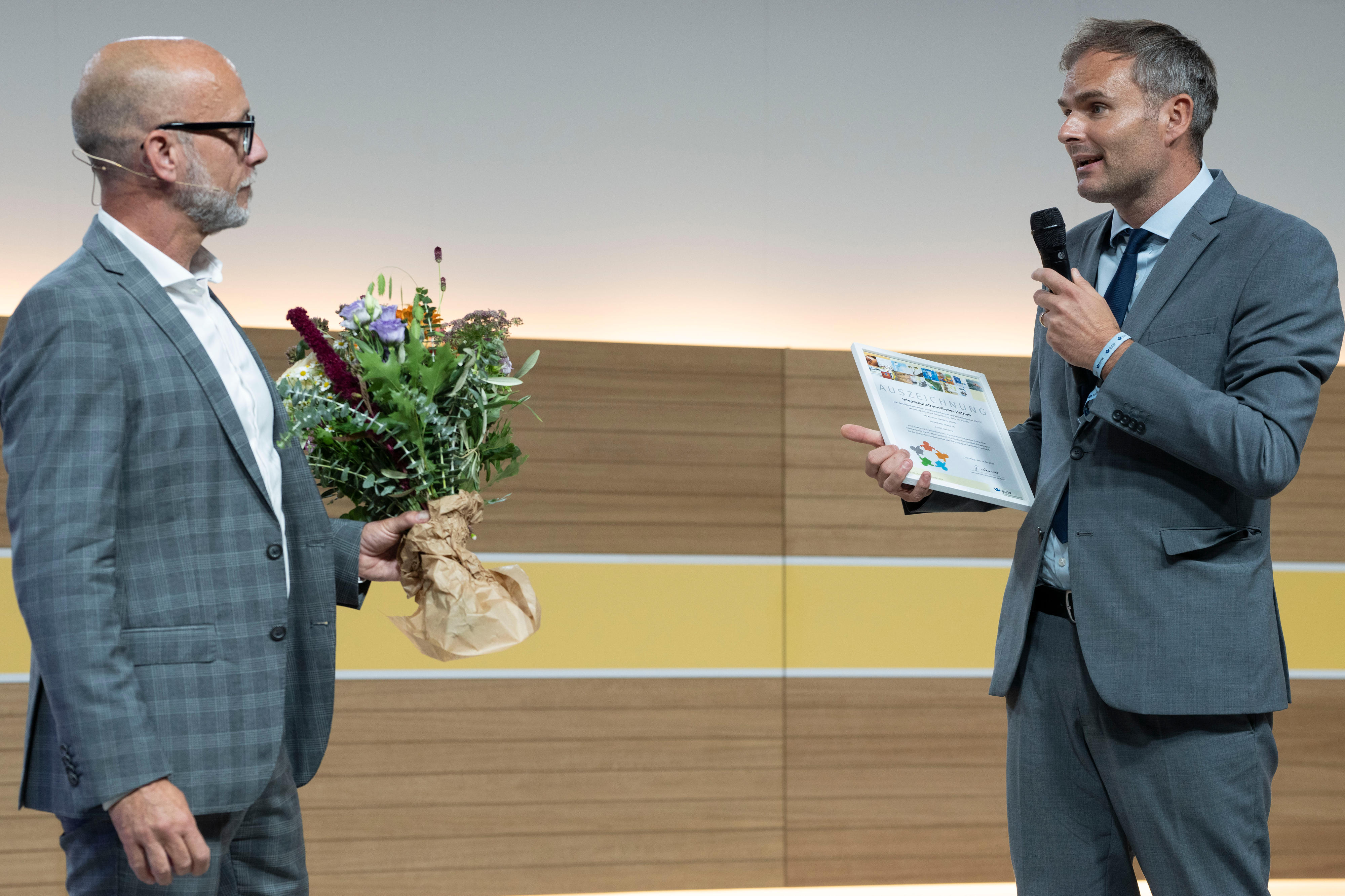 Zwei Männer in Anzug stehen einander zugewwandt auf einer Bühne, der Linke hat einen Blumenstrauß in der Hand, der rechte spricht in ein Mikrofon und hat eine Urkunde in der Hand