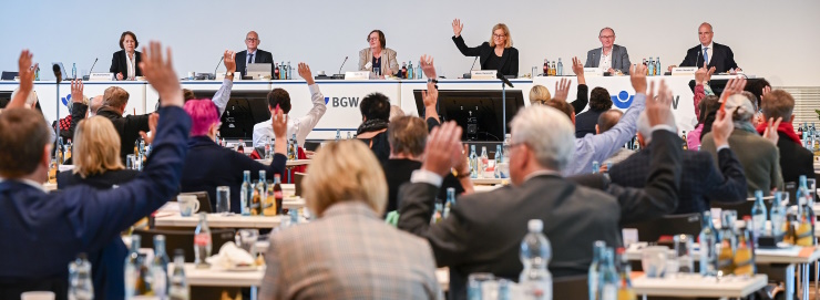 Mitglieder der BGW-Verstreterversammlung sitzen in mehreren Reihen an Tischen, auf denen Getränkeflaschen stehen, und stimmen per Handzeichen ab, und man sieht sie von hinten. Im Hintergrund: das Podium mit weiteren sechs Personen, von denen eine die Hand zur Abstimmung hebt.