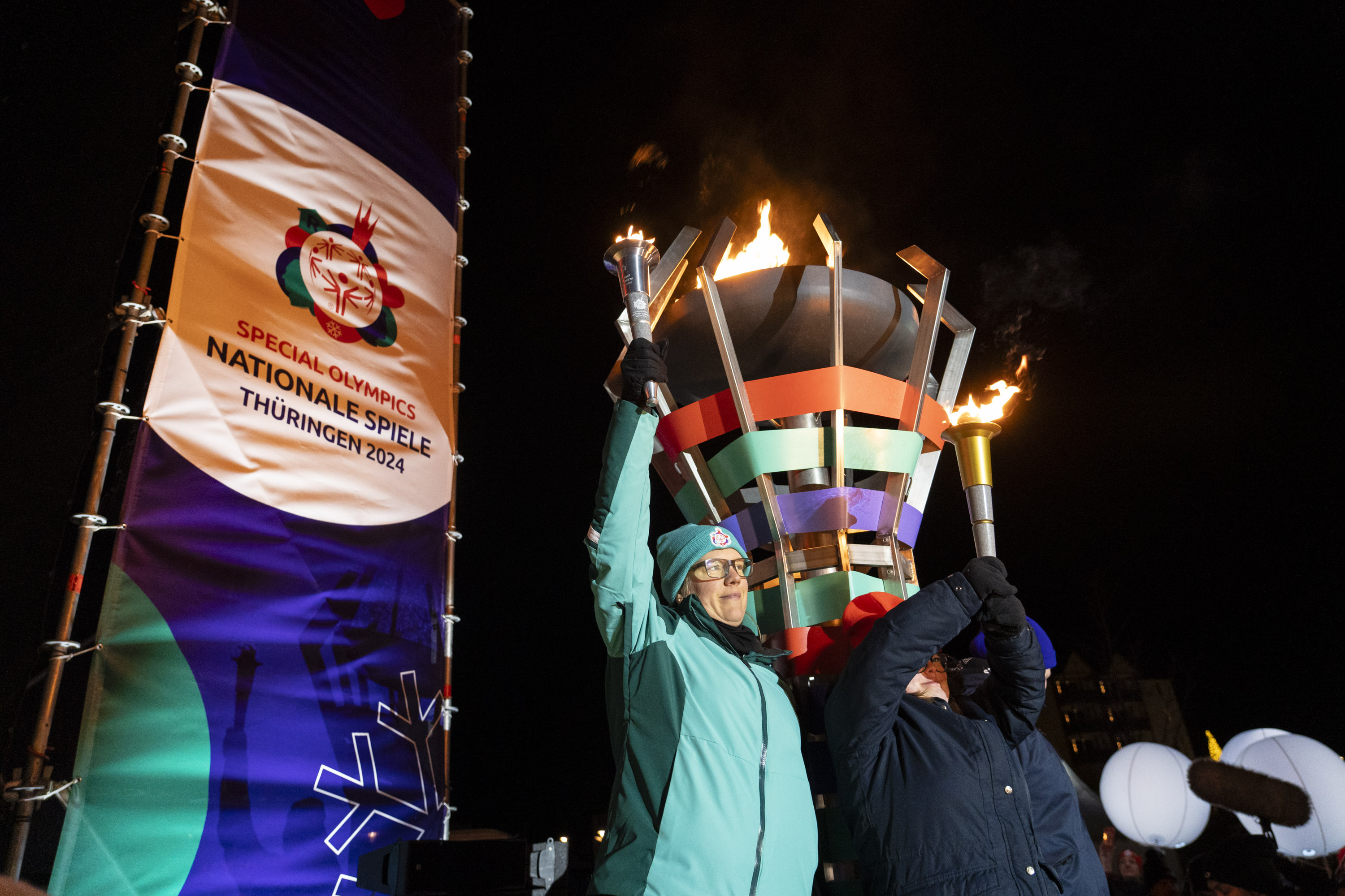 Zwei Sportlerinnen stehen vor der entzündeten Flamme und halten beide Fackeln in der Hand. Links daneben ein Banner mit dem Logo der Spiele.