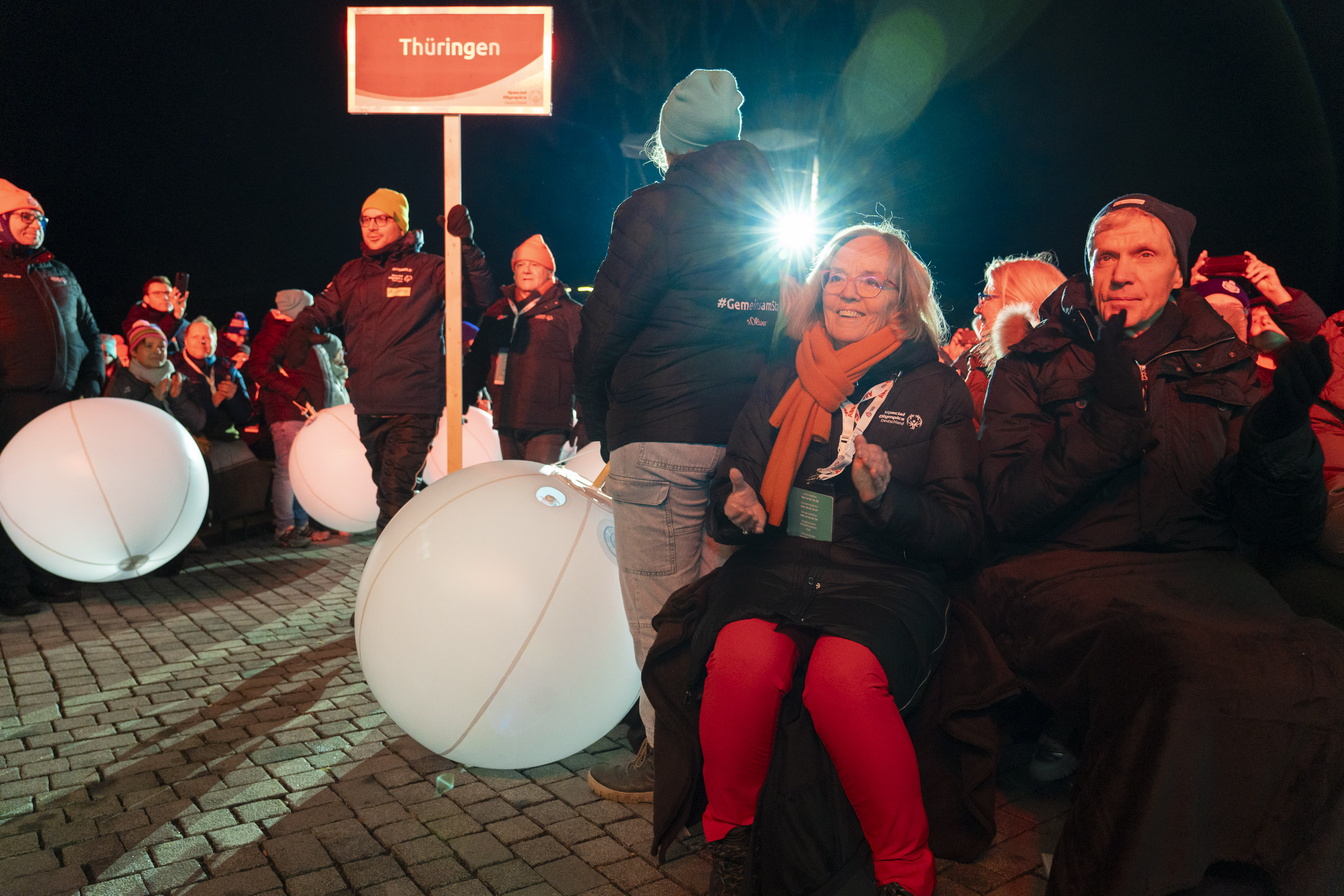 Im Vordergrund sitzen die SOD-Präsidentin und der Sportminister. Links daneben laufen zwei Athleten mit einem Schild "Thüringen" durch die Menge. Helfende am Rand halten große, weiße Bälle.
