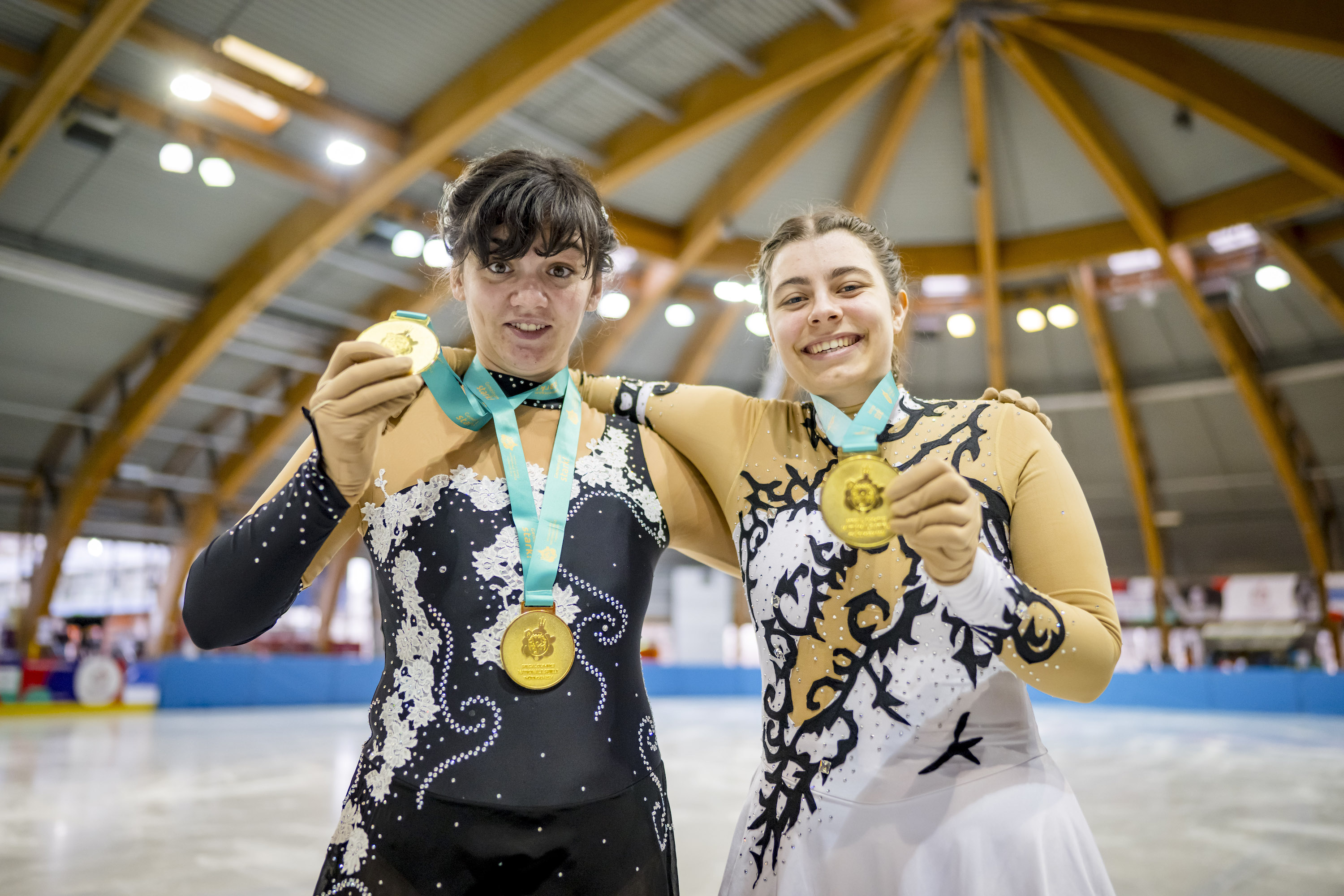 Zwei Frauen stehen nebeneinander in einer Eishalle. Beide tragen ärmellose, dekorative Sportbekleidung und halten stolz ihre gewonnenen Medaillen hoch.