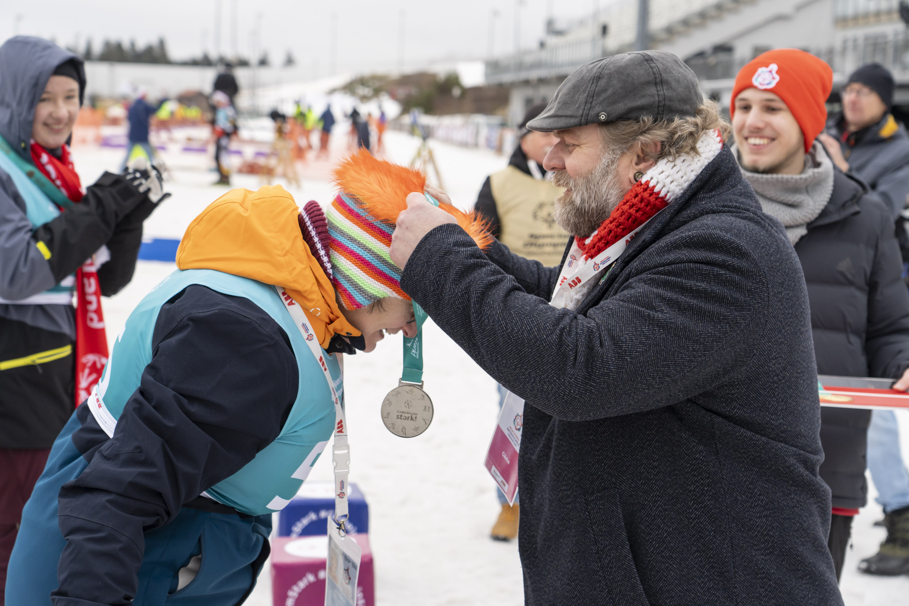 Ein Mann mit Mütze und Winterjacke hängt einer Athletin mit Mütze und Winterjacke eine Silbermedaille um. Im Hintergrund weitere Personen.
