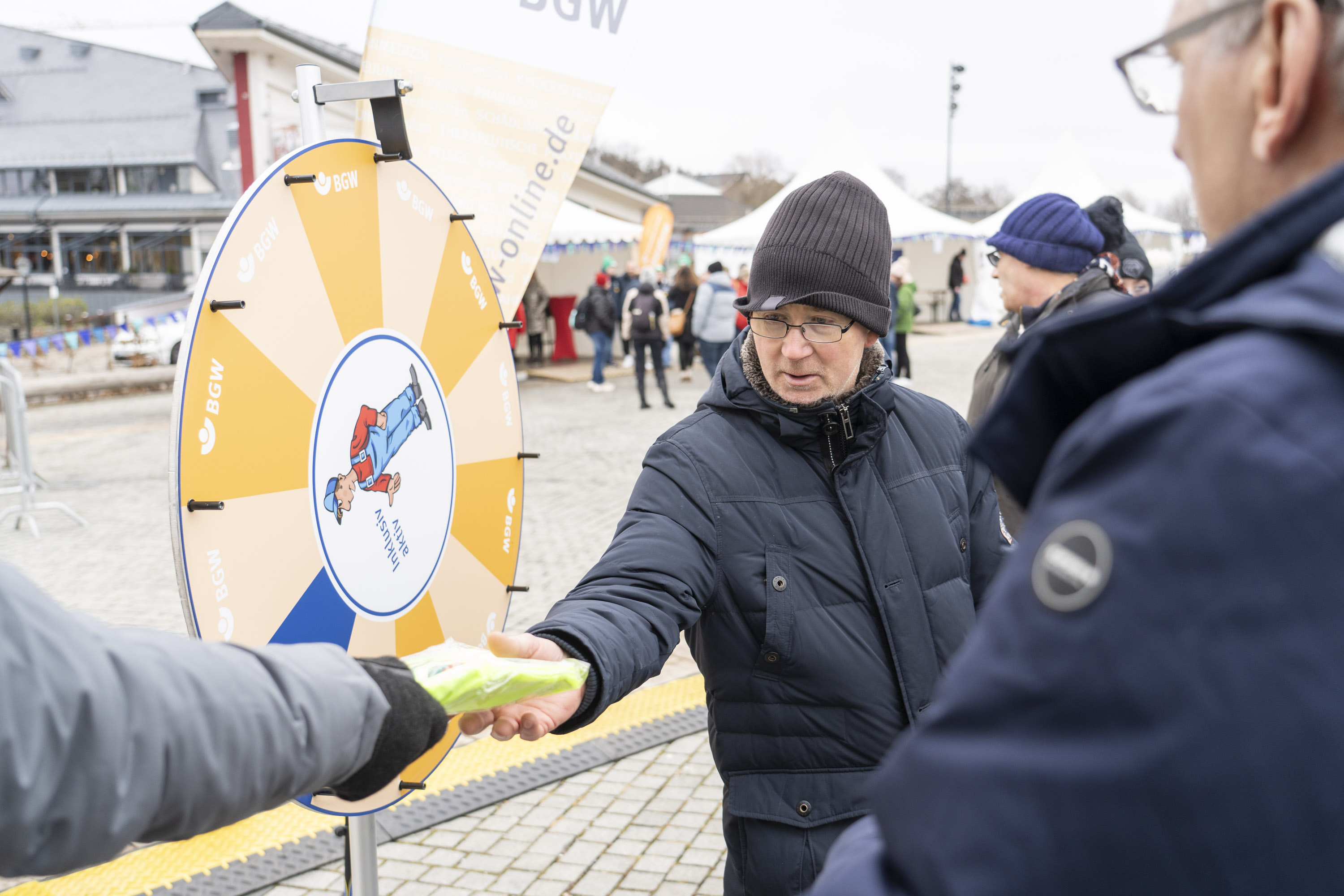 3 Personen vor dem BGW-Glücksrad mit der Figur Nils drauf. Ein Arm reicht der Person in der Mitte einen BGW-Gewinn.