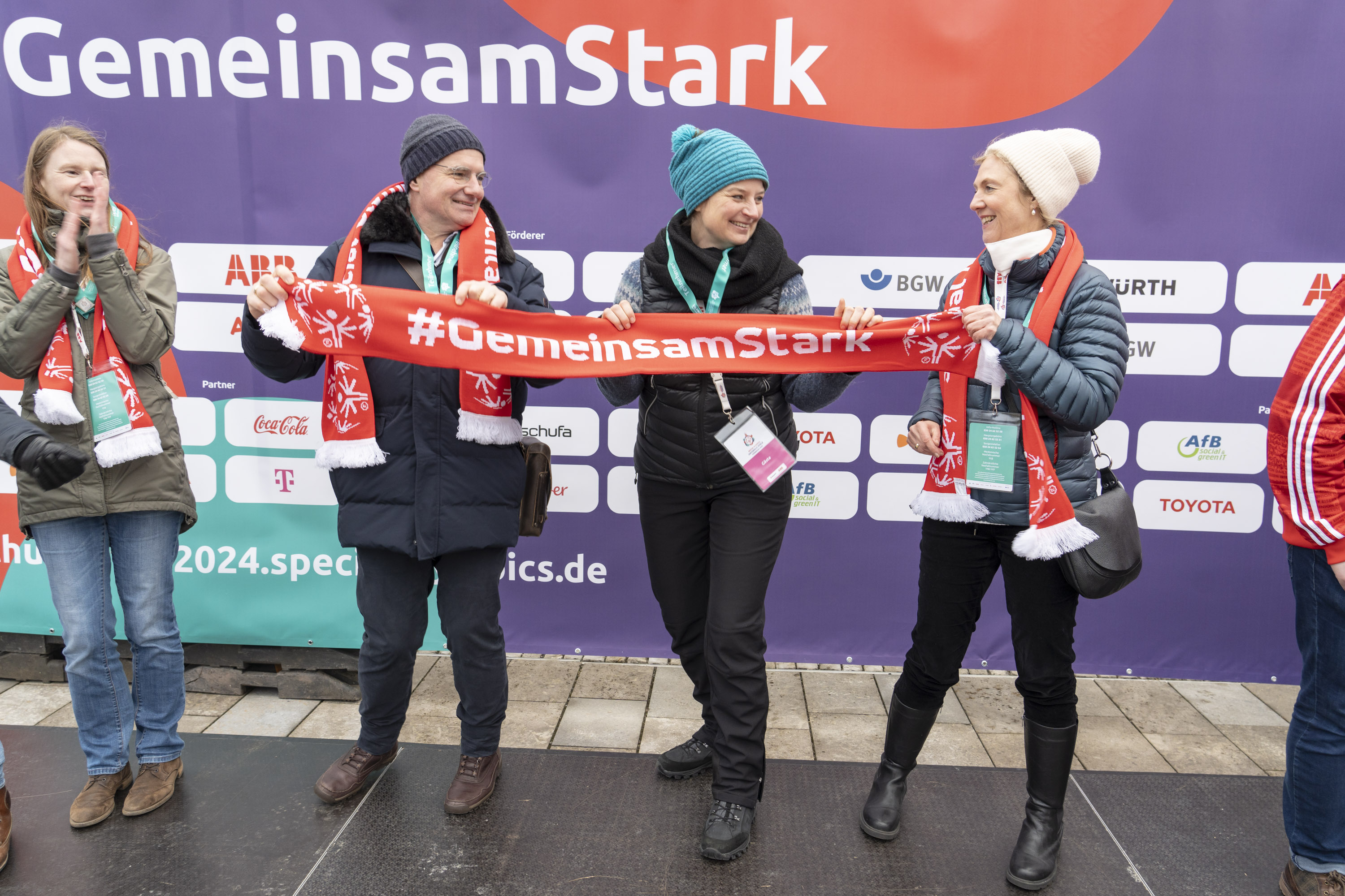 4 Frauen nebeneinander vor einer Wand mit diversen Firmen-Logos und dem Slogan "GemeinsamStark". 3 der Frauen halten zusammen einen Schal mit der Aufschrift "#GemeinsamStark".