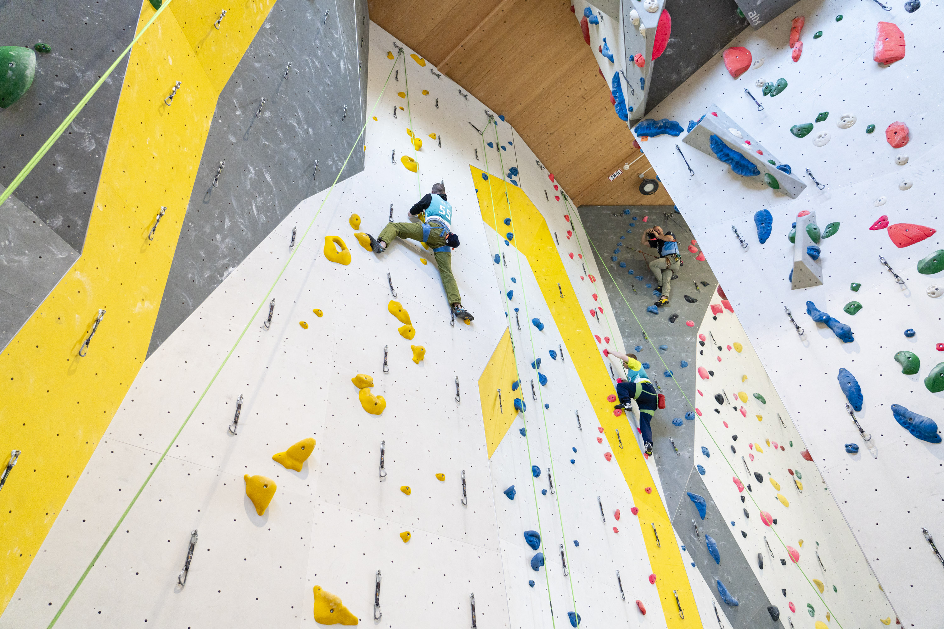 Eine Kletterhalle. Man sieht die Kletterwände mit Routen. 3 Personen klettern an den Wänden.