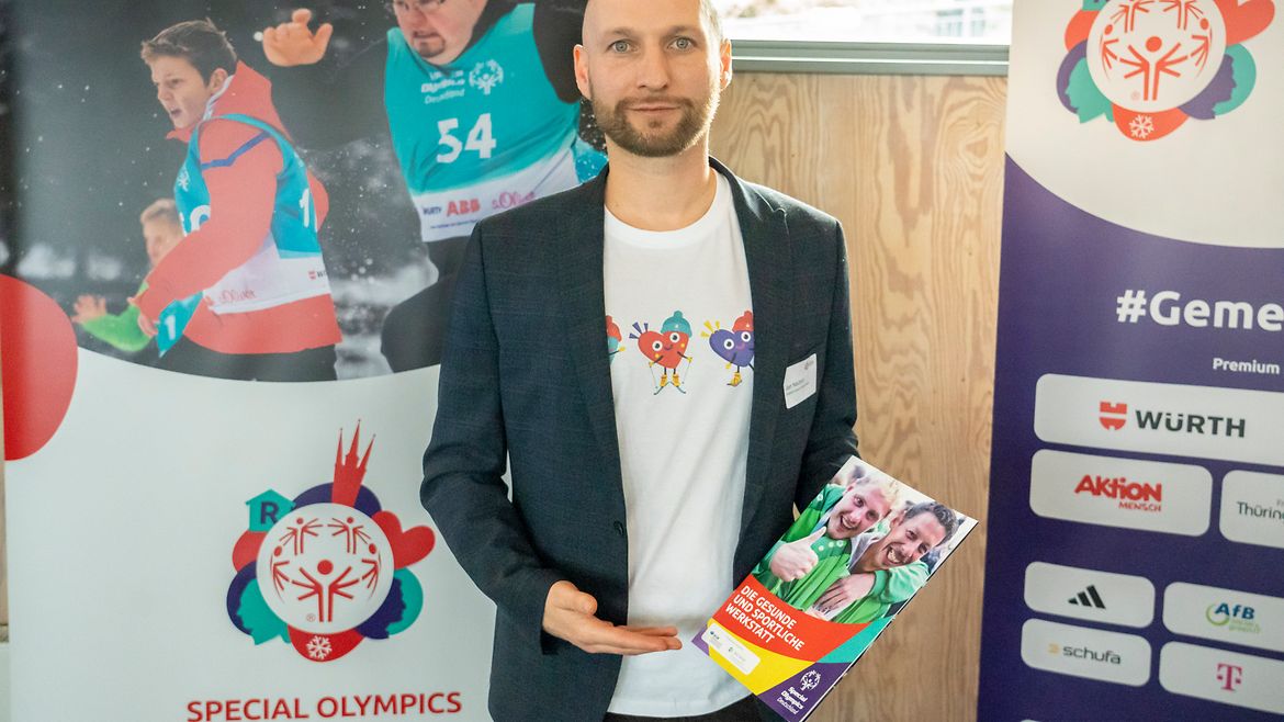 Eni Mann steht vor 2 Plakaten mit SOD-Motiven undverschiedenen Logos. In der Hand hält er die Broschüre "Die gesunde uund sportliche Werkstatt".