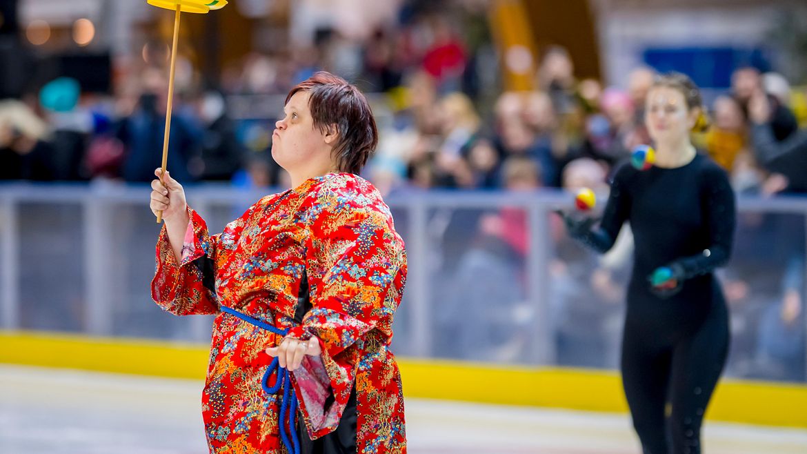 Zwei Athletinnen auf einer Eisfläche im Eisstadion. Eine balanciert einen Teller auf einem Stock, die andere jongliert mit 3 Bällen.