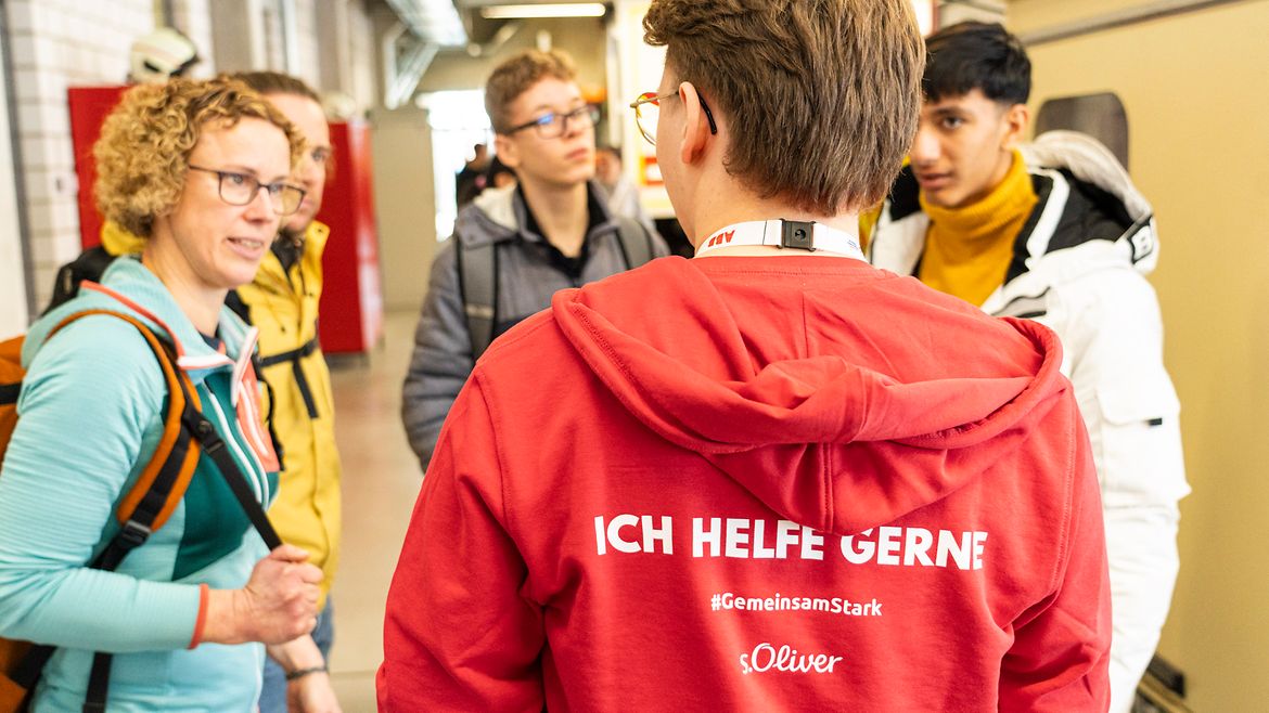 Ein junger Mann von hinten. Er trägt einen roten Pulli, auf dem steht "Ich helfe gerne!" 4 Personen stehen vor ihm und sprechen mit ihm.