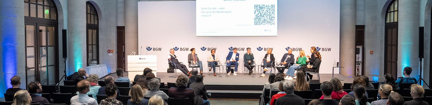 BGW Sportsymposium 2024 10 Menschen mit und ohne Behinderung sitzen auf einer Bühne. Über ihnen eine Leinwand mit "BGW-Sportsymposium, 13. März 2024" und QR-Code. Im Saal davor Publikum auf Stühlen.