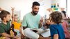 Eine Gruppe Kinder sitzt mit einem Erzieher auf dem Boden. Er hält ein Buch auf dem Schoß, die Kinder strecken die Hände nach oben und melden sich.