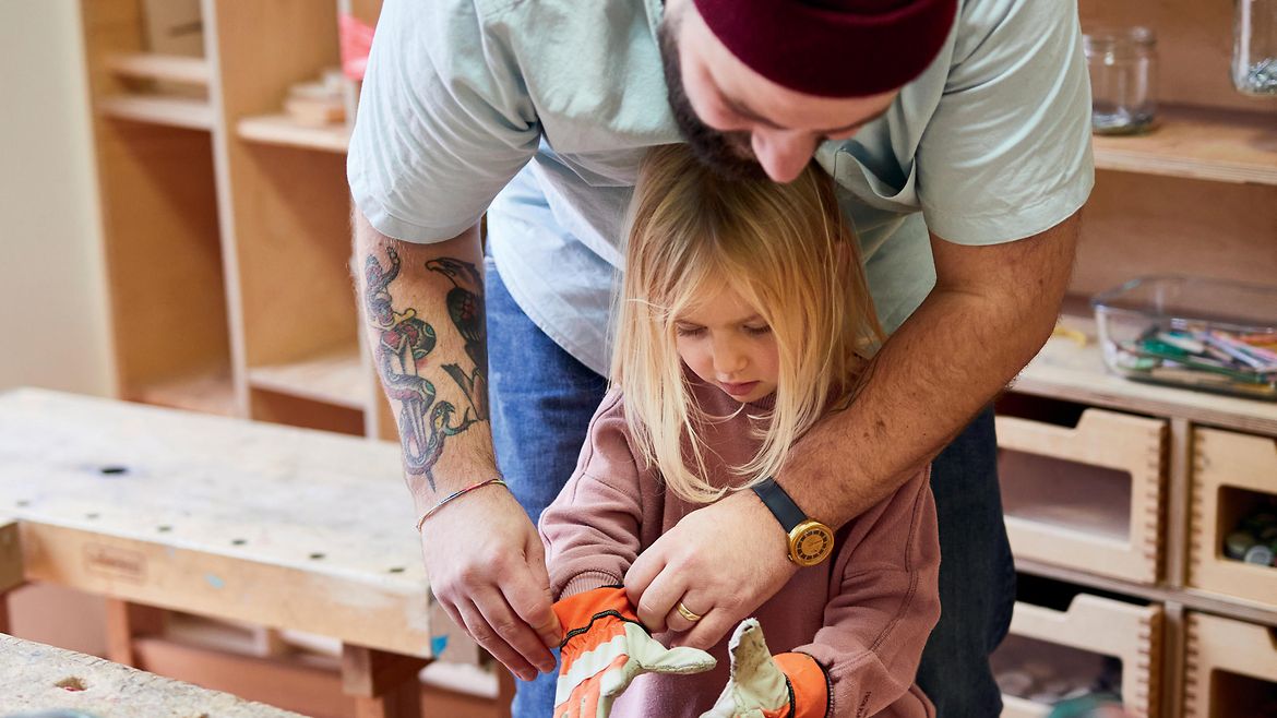 Ein bärtiger Mann mit Wollmütze und Tattoo am Arm hilft einem kleinen Mädchen Arbeitshandschuhe anzuziehen.
