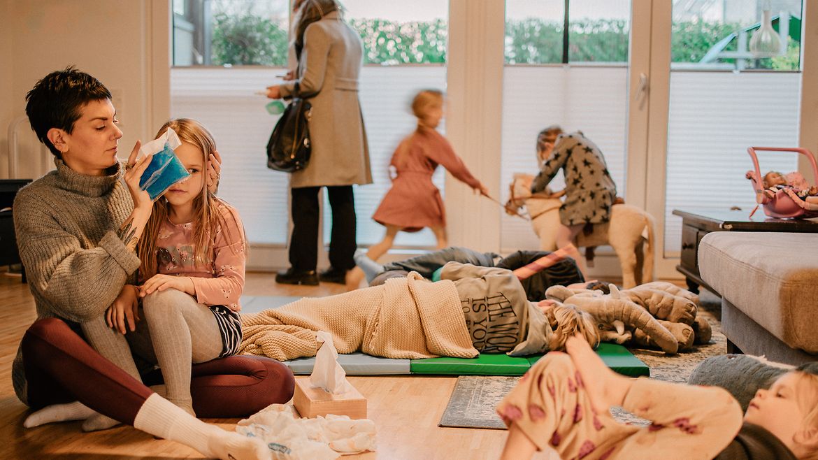 Eine Frau sitzt auf dem Fußboden mit einem Mädchen auf dem Schoß, um sie herum liegen und spielen Kinder