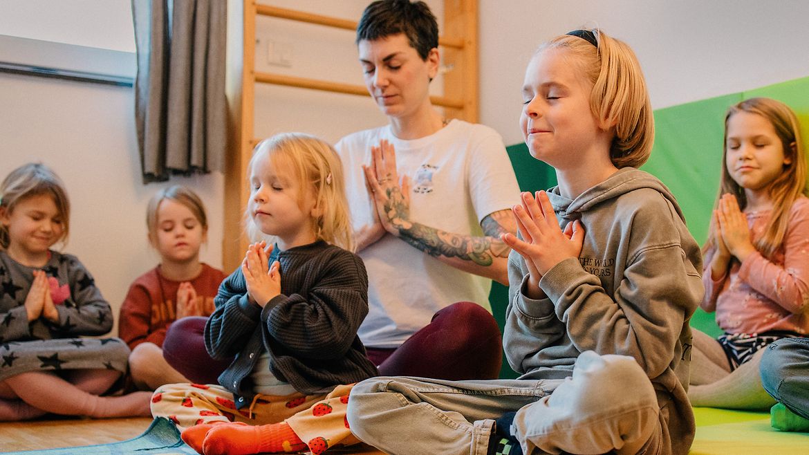 Eine Frau und mehrere Kinder um sie herum sitzen im Schneidersitz auf dem Boden, haben die Hände vor der Brust gefaltet und die Augen geschlossen