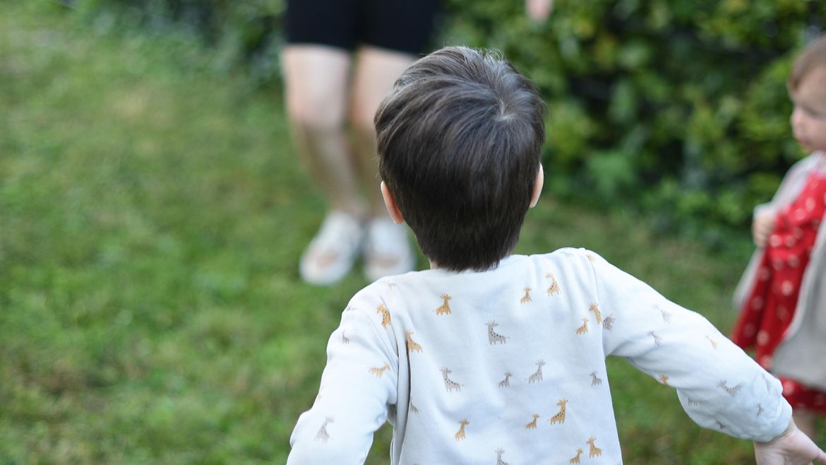 Rückansicht eines Jungen, der auf dem Rasen springt, im Hintergrund ist ein anderes Kind und die beine einer Frau zu erkennen