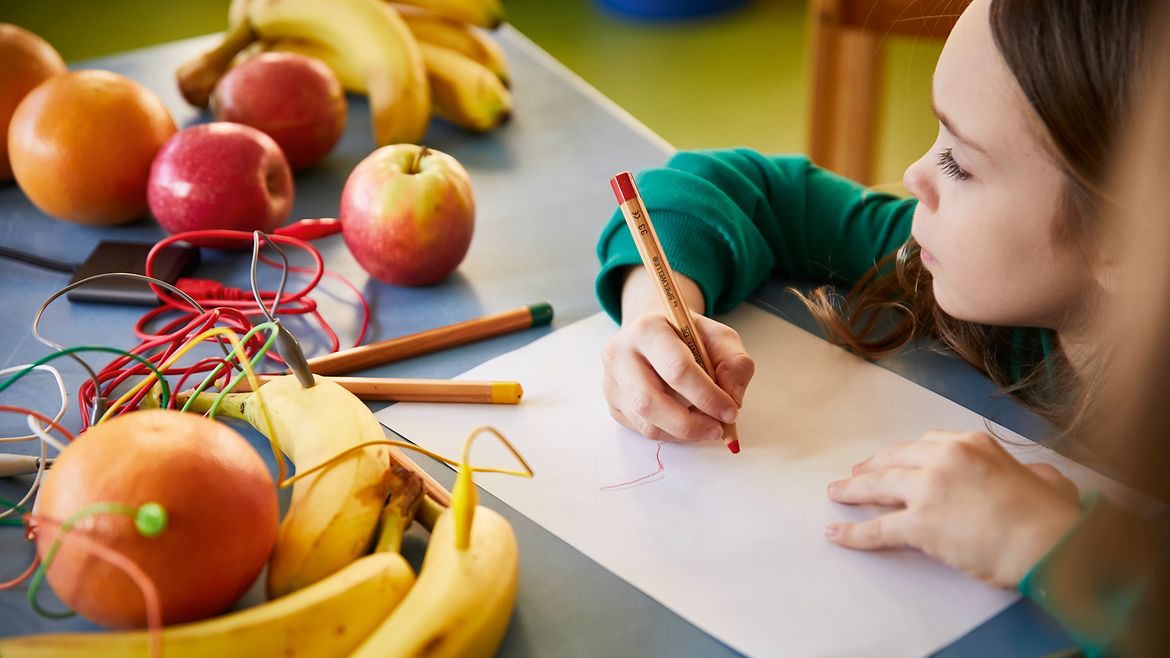 Ein Mädchen sitzt an einem Tisch vor mit Kabeln verbundenen Äpfeln und Bananen und malt.