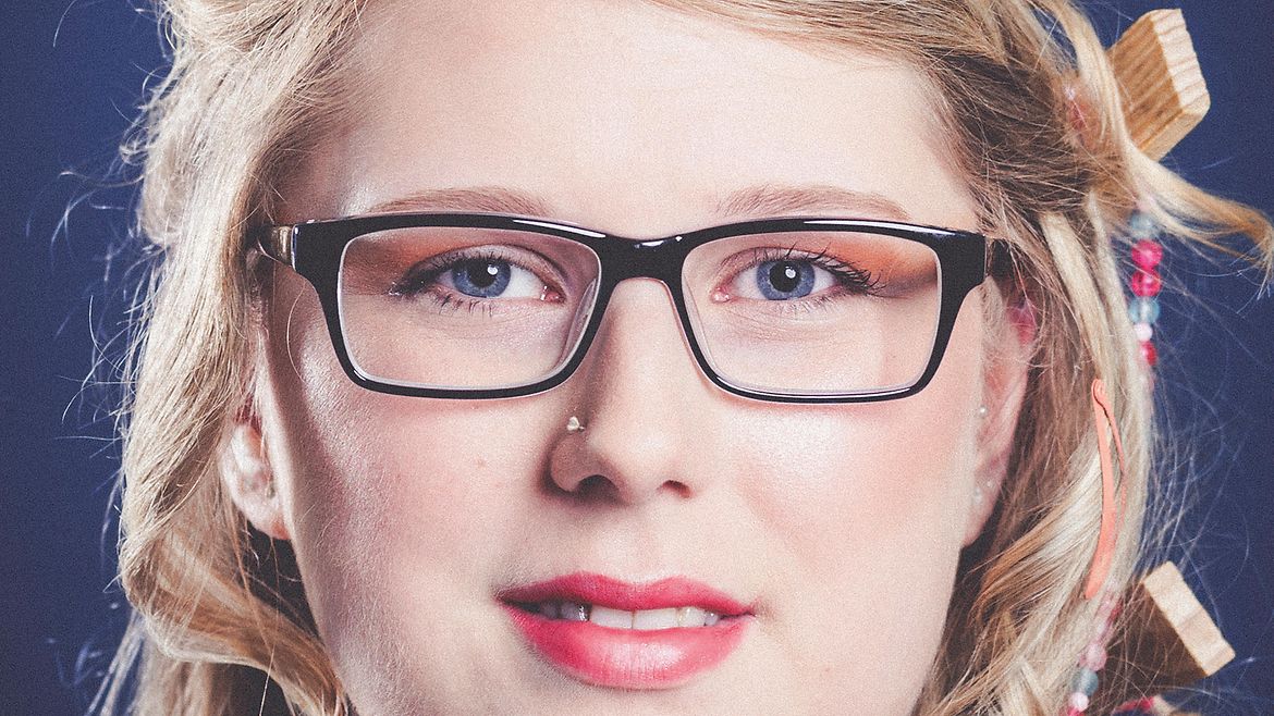 Porträt einer blonden, lächelnden Frau mit Brille mit bunten Spangen und Holzscheiben im Haar
