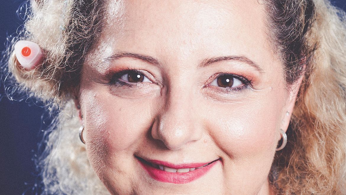 Porträt einer blonden, lächelnden Frau mit bunten Stiften im Haar