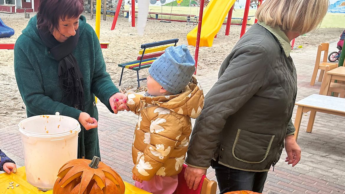 Eine Frau steht mit einem Kind an einem Tisch, auf dem zwei Kürbisse stehen, und lacht, während eine andere Frau sich vom Tisch abwendet.