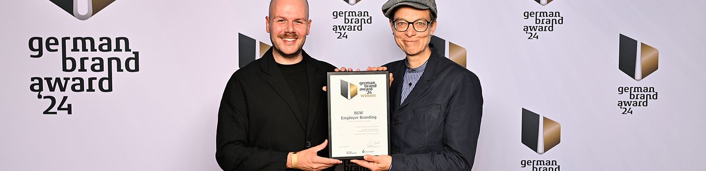 André Holonics und Tanja Rieckmann-Mekhchoun halten gemeinsam eine gerahmte Urkunde hoch vor einer weißen Wand mit mehreren Logos des German Brand Award 24