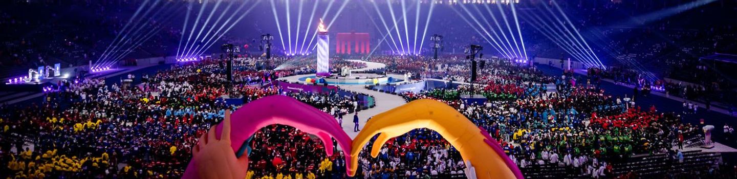 Eröffnungsfeier SOWG 2023 im Berliner Olympiastadion Eröffnungsfeier der Weltspiele im Berliner Olympiastadion: Scheinwerfer und lila Beleuchtung, in der Mitte ein Meer aus Menschen, im Vordergrund zwei Luftschlangenhände, die sich berühren.