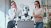 Ein Roboter sitzt mit Headset vor einem Laptop am Schreibtisch in einem Büro. Dahinter stehen ein Mann und eine Frau, die ihn skeptisch bis