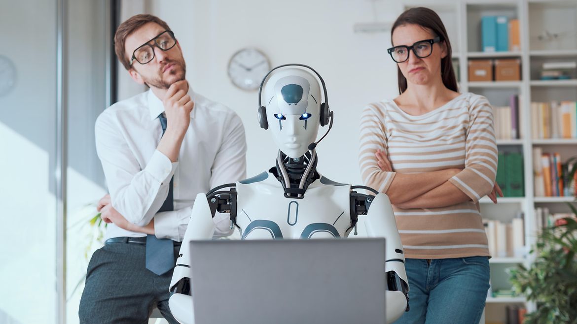 Künstliche Intelligenz Ein Roboter sitzt mit Headset vor einem Laptop am Schreibtisch in einem Büro. Dahinter stehen ein Mann und eine Frau, die ihn skeptisch bis