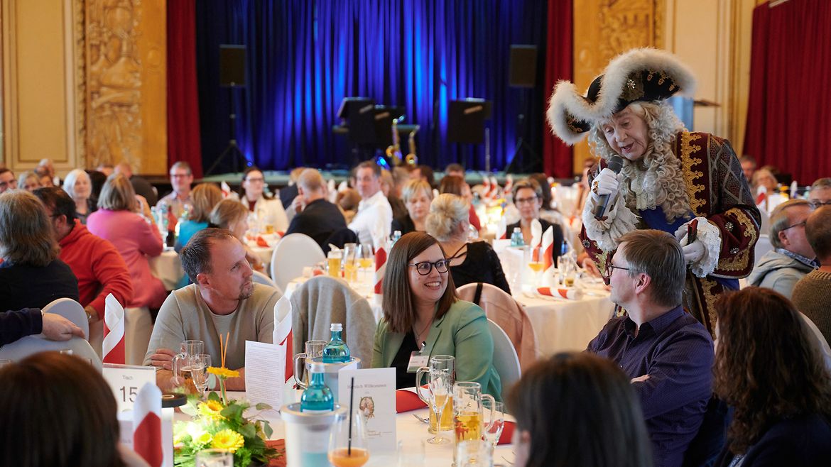 Auf einer Abendveranstaltung steht ein Mann in einem aufwändigen Barockkostüm mit Hut und Perücke an einem Tisch, an dem Personen sitzen und spricht in ein Mikrofon.
