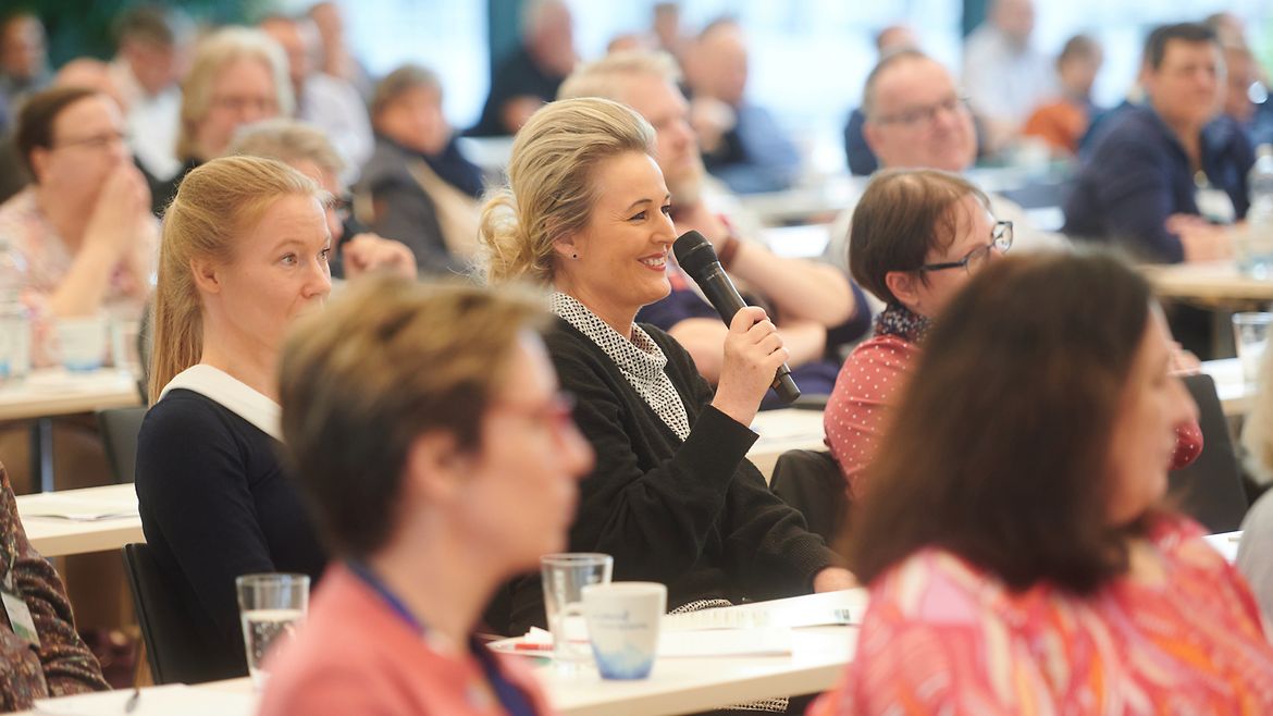 Im Kongress-Publikum shält eine blonde Frau ein Mikrofon in der Hand und lächelt.