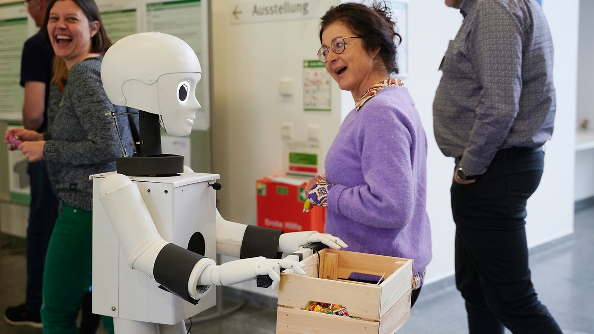 Ein Roboter mit großen Augen und lächelndem Gesicht hält eine Holzkiste mit bunten Gegenständen in den Händen. Zwei Frauen gehen vorbei und schauen ihn lachend an.