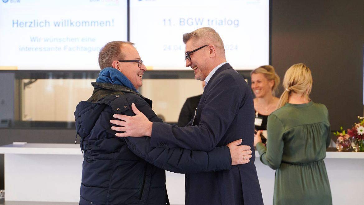 Zwei Männer begrüßen sich herzlich, im Hintergrund ist ein Tresen und Monitore auf denen steht "Herzlich wilkommen" und "11. BGW trialog".