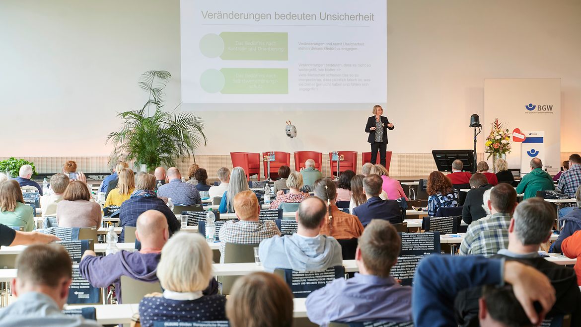 Eine Frau steht auf einer Bühne vor sitzendem Publikum. Hinter Ihr ist eine Präsentation an die Wand projiziert mit dem Text "Veränderungen bedeuten Unsicherheit".