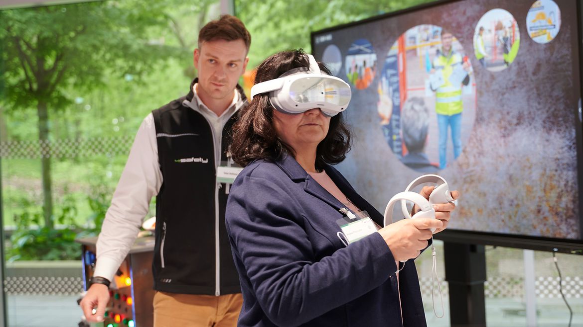 Eine Frau mit VR-Brille auf dem Kopf und zwei Controllern in den Händen steht vor einem Bildschirm.