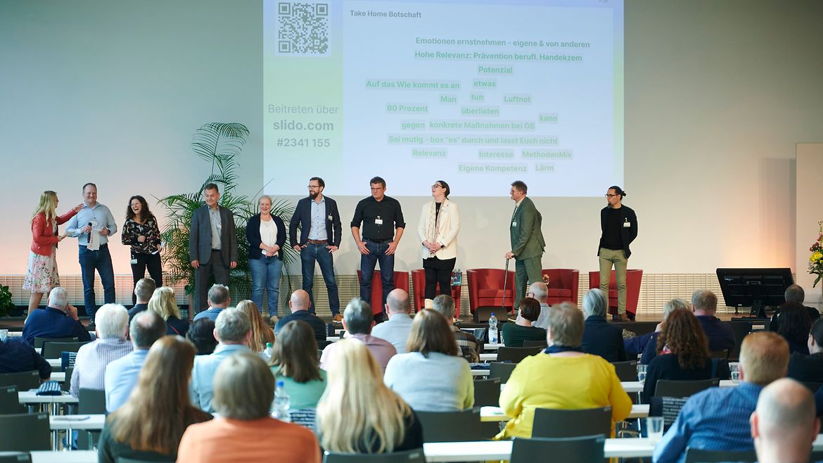 Zehn Personen stehen auf einer Bühne vor sitzendem Publikum, im Hintergrund ist eine Präsentation an die Wand projeziert.