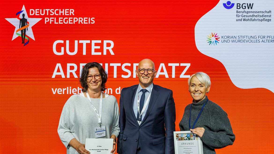 Jörg Schudmann, Hauptgeschäftsführer der BGW, zusammen mit Marita Frerichs und Stefanie Freimuth-Hunfeld, die die Urkunde hält, vom St. Nikolausstift Caritas Pflege aus Papenburg auf der Bühne. .