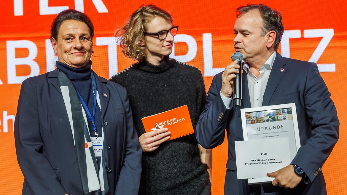Der Moderator zusammen mit Heidrun Grunwald und Thomas Kraska, der in ein Mikrofon spricht und die Urkunde hält, von den DRK Kliniken Berlin Pflege und Wohnen Mariendorf auf der Bühne.