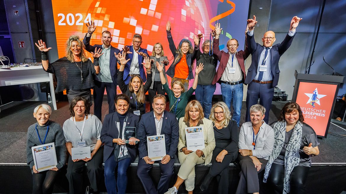 18 Personen, die auf einer Bühnenkonstruktion stehen und sitzen, einige davon halten die Hände in die Höhe. Im Hintergrund ist eine Leinwand, auf der eine Dissokugel zu sehen ist und die Aufschrift 2025, rechts im Bild ein Rednerpult mit der Aufschrift Deutscher Pflegepreis 2024.